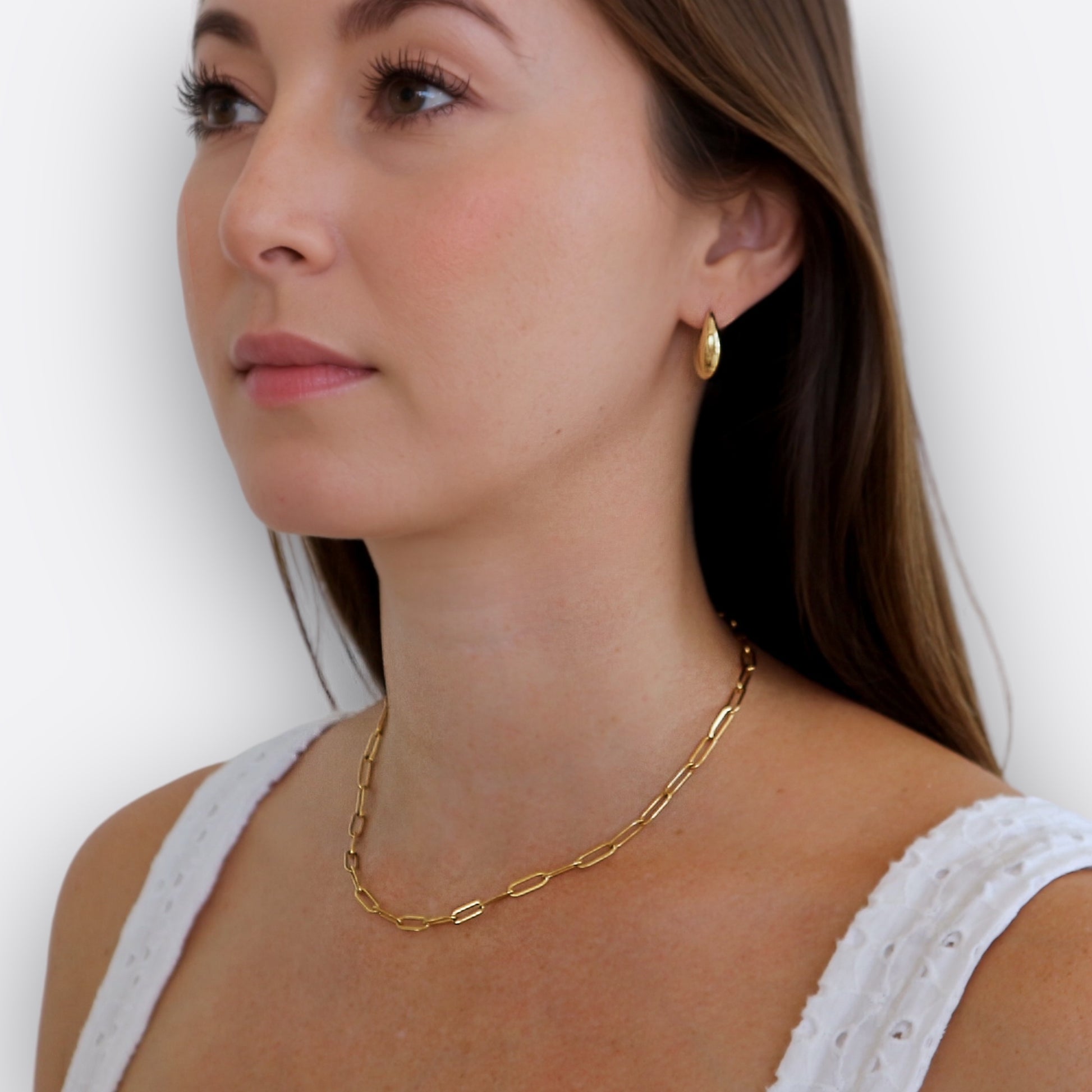 brunette girl showing her profile wearing gold earrigs, paperclip necklace and a white tank. white background
